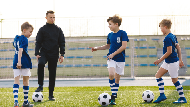 Treneris futbola laukumā kopā ar 2 zēniem un futbola bumbām