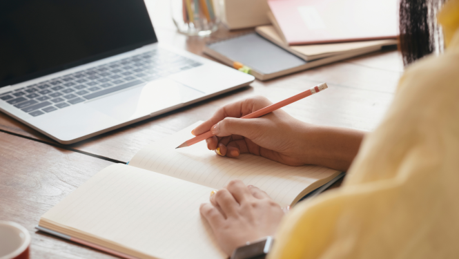 Sievietes rokas tur zīmuli un raksta kladē, uz galda dators 