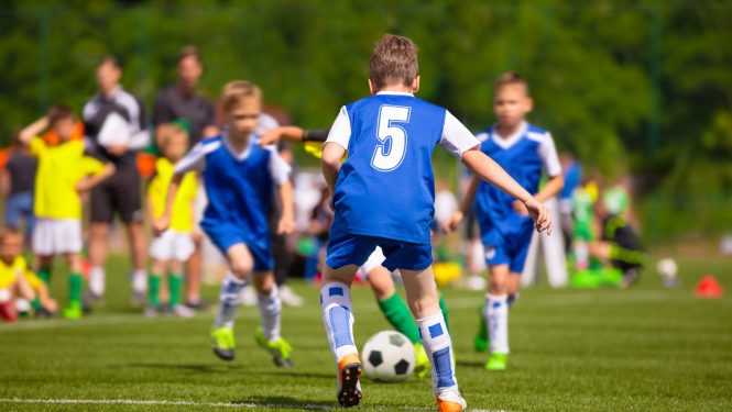 Puikas zilās formās spēlē futbolu. Fonā stāv citi puikas dzeltenās formās un treneri.