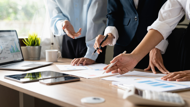 Darba galds ofisā, uz kura ir dažādi papīri un cilvēki ar pildspalvām rokās tos pārskata