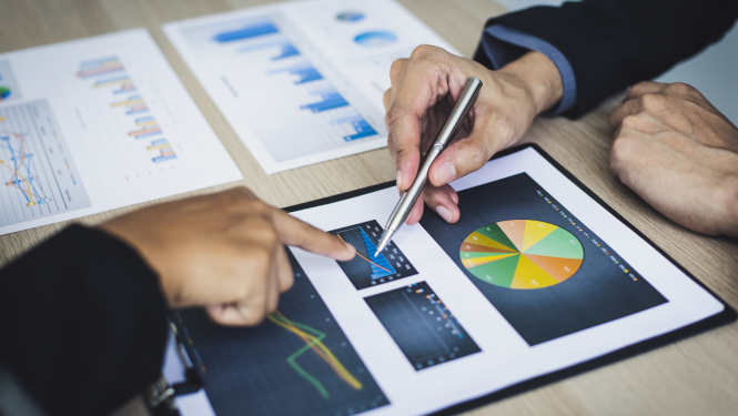 Uz galda noliktas krāsainas diagrammas, divu cilvēku rokas rāda uz vienu no grafikiem