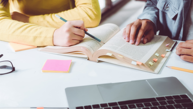 Gramata un dators stāv uz galda, cilvēku rokas velk līdzi tekstam 