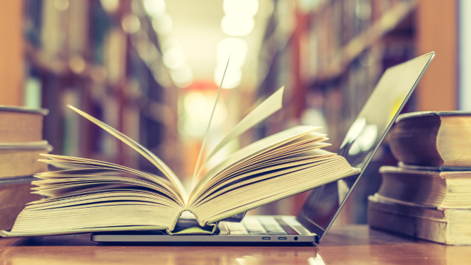 Bibliotēkā uz galda stāv grāmatas un atvērts portatīvais dators, uz kura nolikta atvērta grāmata