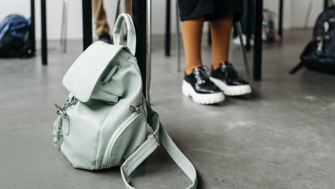 Priekšplānā mugursoma uz grīdas klasē, redzamas bērnu kājas un galdu kājas