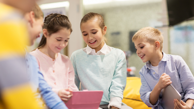 Pieci priecīgi bērni sēž kopā, skatās planšetē, fokusā divas meitenes un puika
