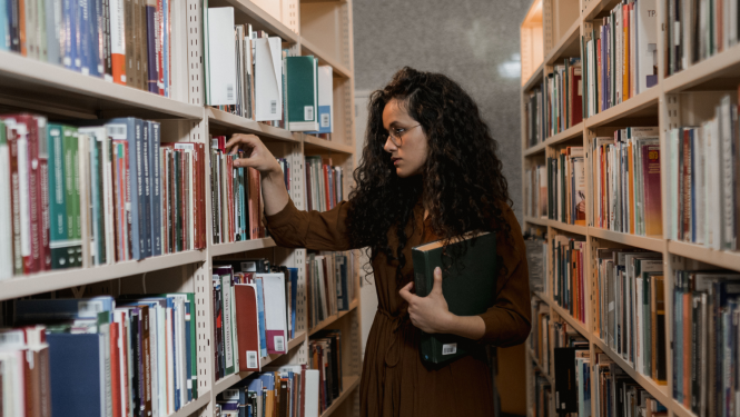 Tumšmataina sieviete  ar grāmatām rokās meklē grāmatas bibliotēkā grāmatu plauktā
