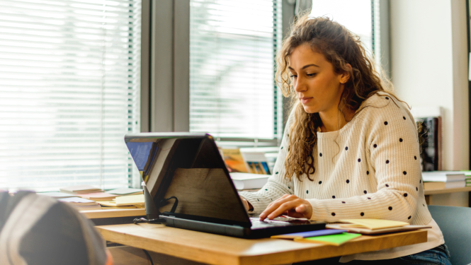 Meitene gaišā džemperī sēž pie galda un raksta klēpjdatorā