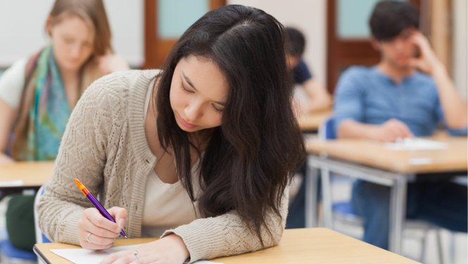 jaunieši klasē raksta eksāmenu, priekšplānā tumšmataina meitene sēž pie galda un raksta uz lapas, roku nolikusi uz lapas