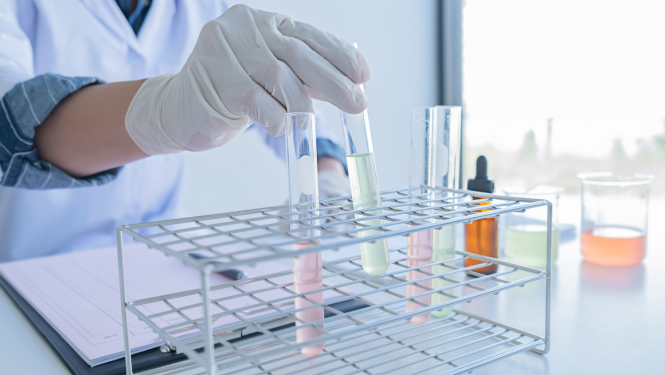 Cilvēka roka baltos gumijas cimdos liek statīvā mēģenes ar gaiši rozā un zaļganiem šķīdumiem