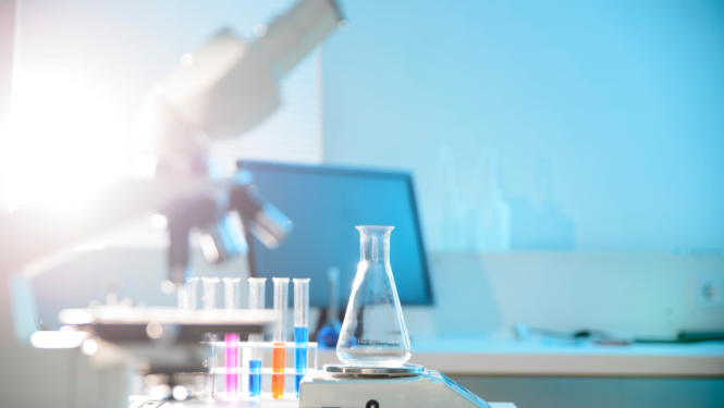 Saules apspīdētā laboratorijā uz galda stāv mikroskops, kolbas un mēģenes ar krāsainiem šķīdumiem, fonā ir datora ekrāns