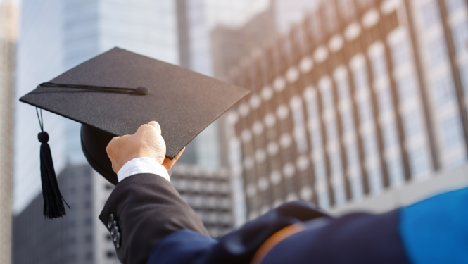 Cilvēka roka tur universitātes absolventa cepuri uz debesskrāpju fona