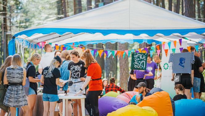 Jaunieši pulcējas pie telts. Ap telti sakārtas krāsainu karodziņu virtenes, blakus novietoti krāsaini sēžammaisi. 