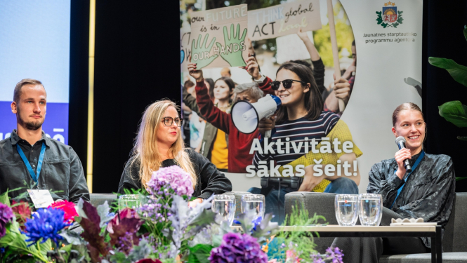 Viens jauns vīrietis un divas jaunas sievietes sēž uz skatuves un runā, priekšplānā ziedu pušķis, aizmugurē plakāts, uz kura attēloti jaunieši un uzraksts "Aktivitāte sākas šeit!"