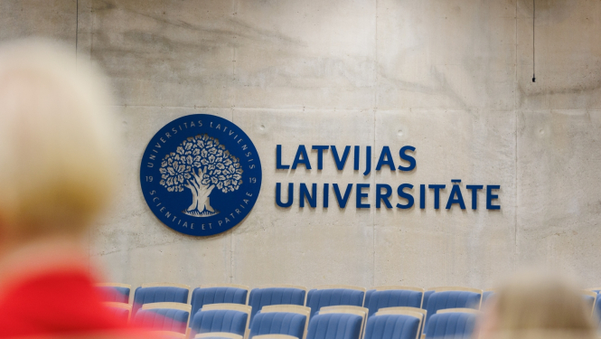 Latvijas Universitātes auditorija ar ziliem krēsliem, uz sienas liels zils LU logotips - ozols zilā aplī un uzraksts Latvijas Universitāte