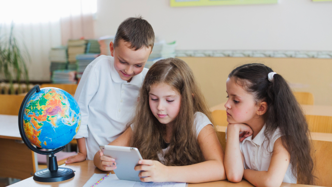 Trīs bērni ar planšeti sēž pie galda, uz galda globuss