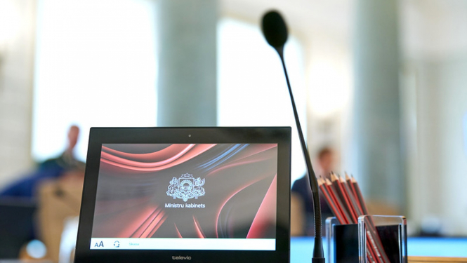 Dators un mikrofons stāv uz galda Ministru kabineta zālē, datora ekrānā redzams Ministru kabineta logotips