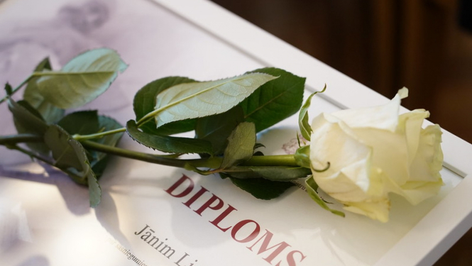 Attēlā redzams Ministru kabineta diploms baltā rāmī un tam virsū uzlikta balta roze
