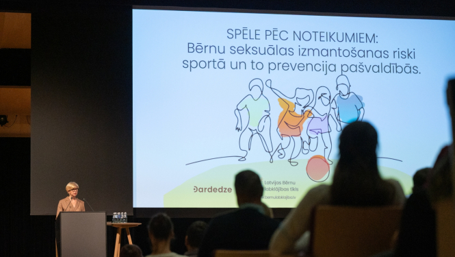 Izglītības un zinātnes ministre Anda Čakša konferencē uz skatuves, aiz viņas liels ekrāns ar bērnu ilustratīvu zīmējumu