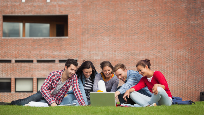 Pieci jaunieši sēž zālājā un skatās portatīvā datora ekrānā, fonā ir sarkanu kieģeļu siena
