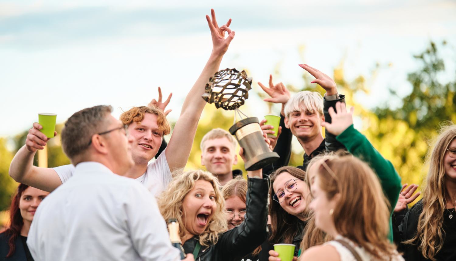 Grupa jaunu cilvēku svin pasākuma norisi brīvā dabā, smaidot un priecājoties. Atmosfēra ir dzīvespriecīga un draudzīga, fonā redzami koki un gaišas debesis.