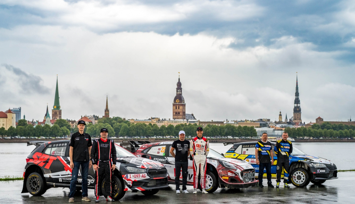 Fonā Rīgas ainava, priekšplānā 3 rallija mašīnas un pie katras stāv 2 sportisti