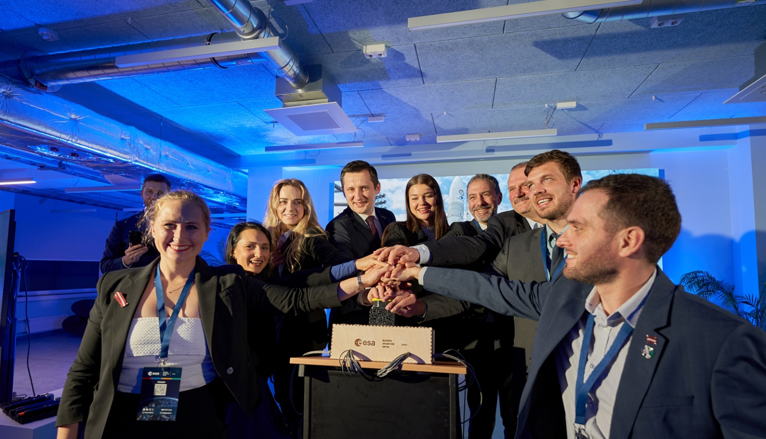 Cilvēki salikuši rokas uz lielas pogas kopīgi to spiež, simboliski palaižot  Eiropas Kosmosa aģentūras Biznesa inkubācijas centra “ESA BIC Latvia” darbību