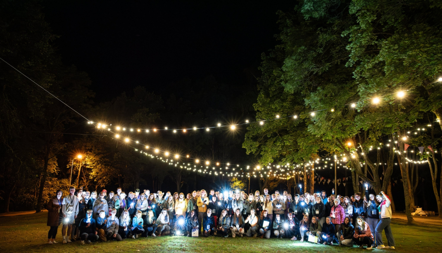 Projektu nakts 2023. dalībnieku kopbilde, vakarā, kad debesis ir tumšas, virs dalībniekiem iekārtas gaimas lampiņu virtenes