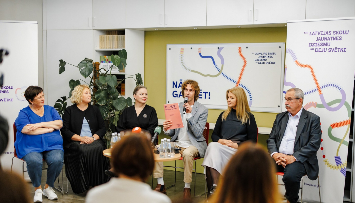 XIII Latvijas Skolu jaunatnes dziesmu un deju svētku preses konference. Skats no zāles, priekšā sēž seši cilvēki un stāsta par pasākuma norisi.