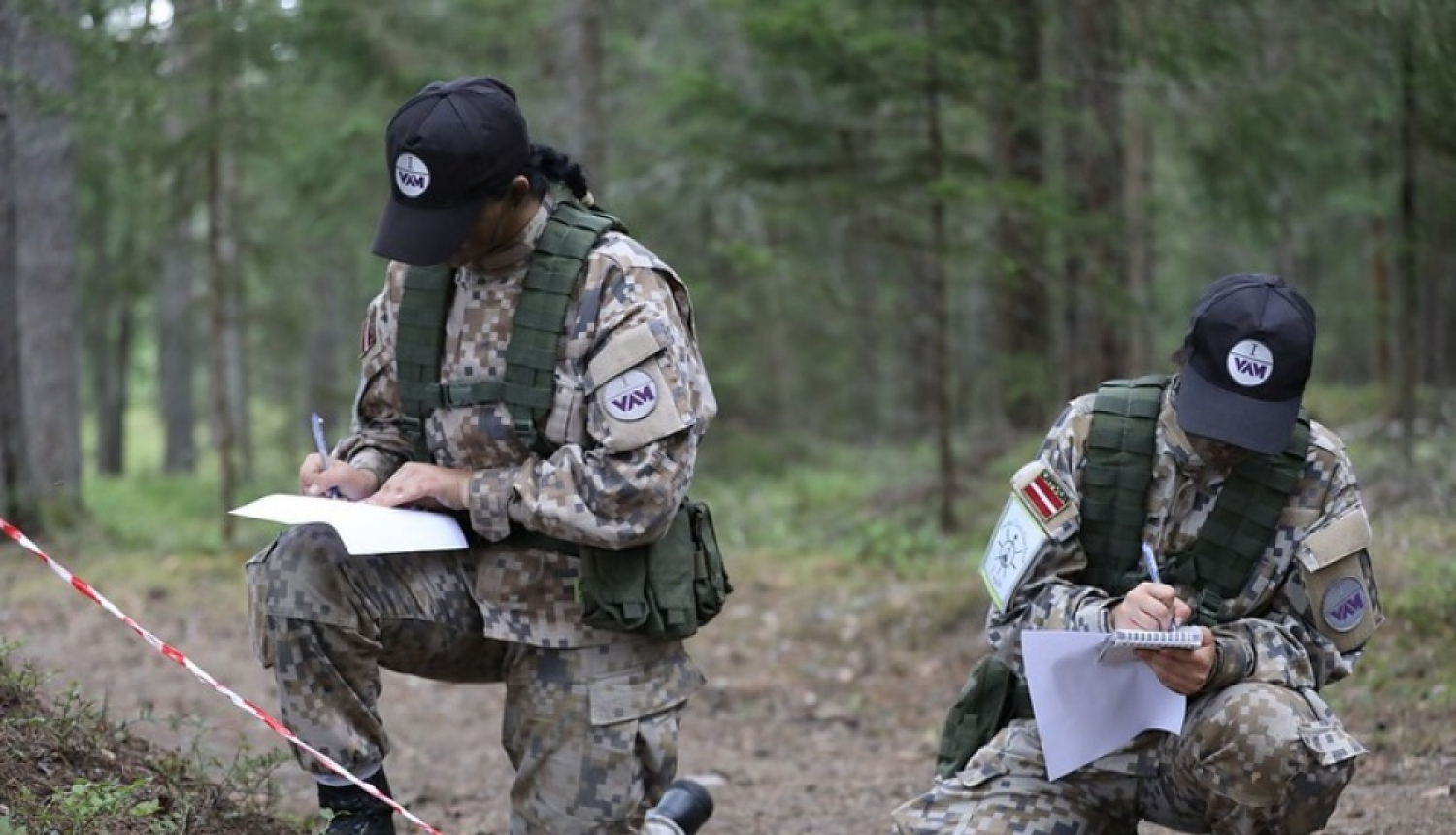 Cilvēki karavīru formas tērpos, veic pierakstus uz lapas dabā