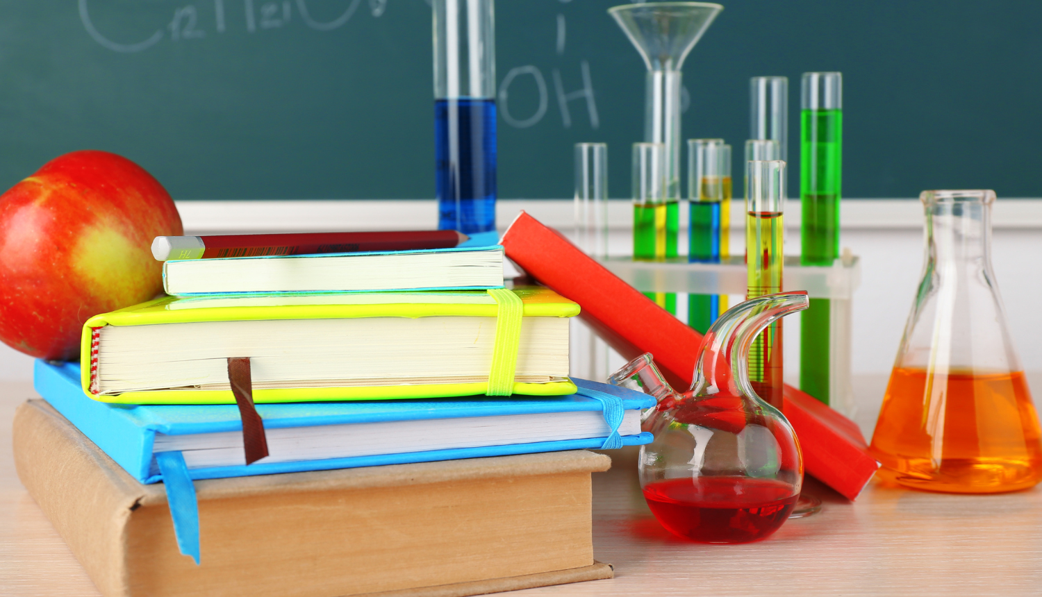 Galds klasē ar grāmatām, ābolu, kolbām, kurās ir dažādu krāsu šķīdumi. Fonā tāfele, uz kuras uzrakstītas ķīmijas formulas.