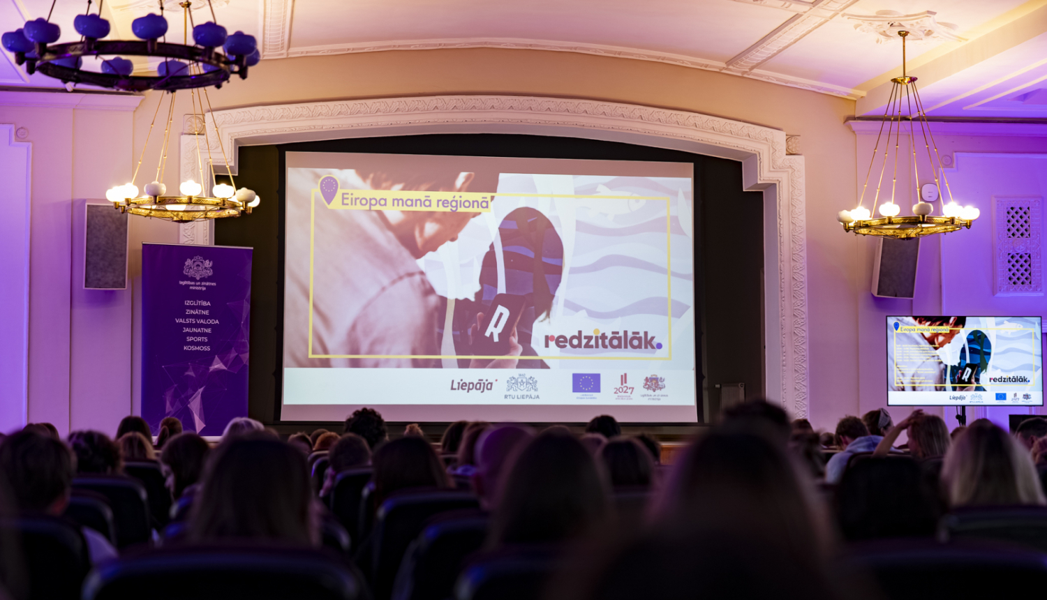 Pasākuma "Eiropa manā skolā" titulbilde uz lielā ekrāna RTU Liepāja lielajā zālē, zāle pilna ar pasākuma dalībniekiem
