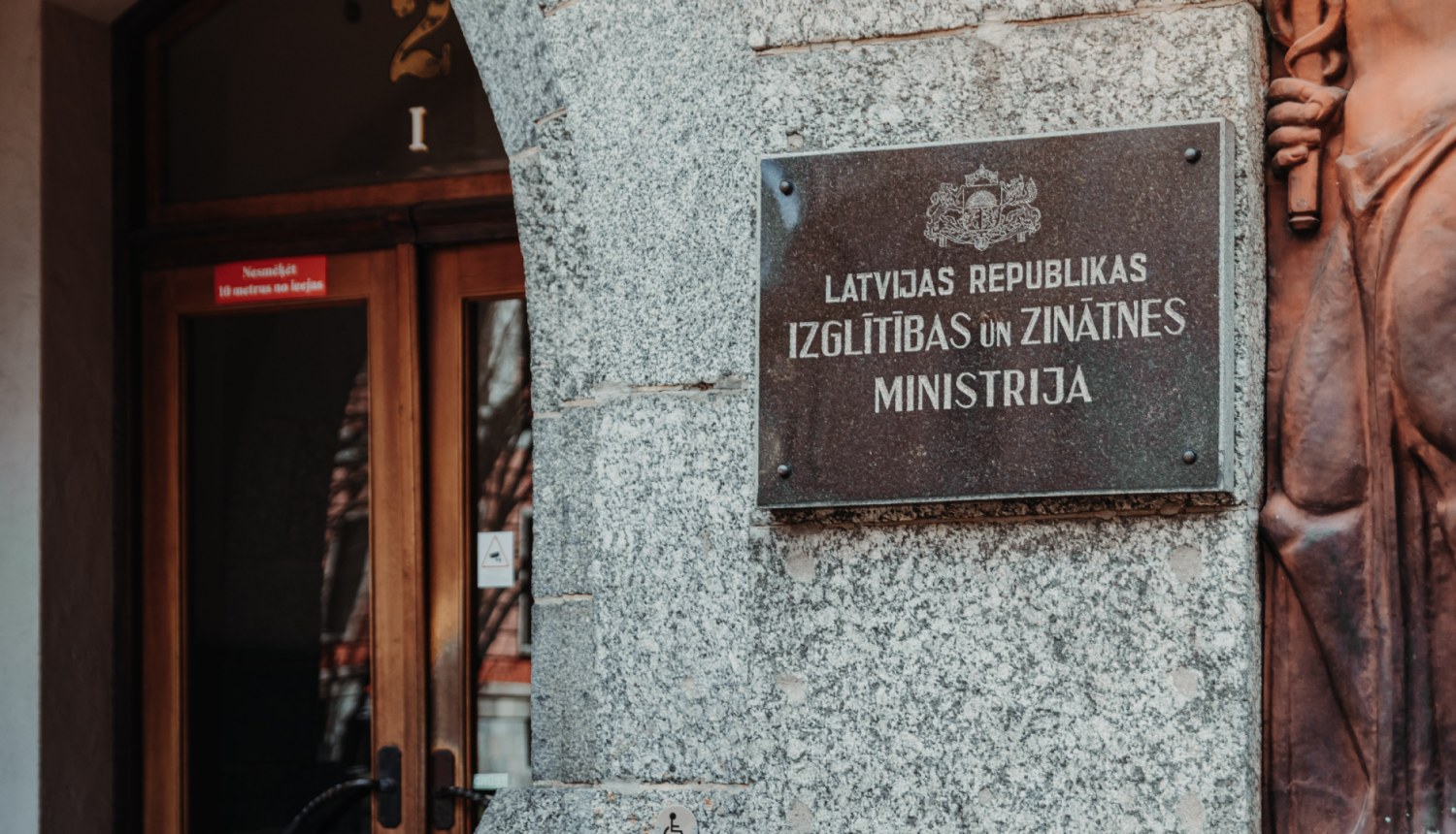 Izglītības un zinātnes ministrijas ieejas durvis, pie sienas plāksne ar IZM logo un uzrakstu