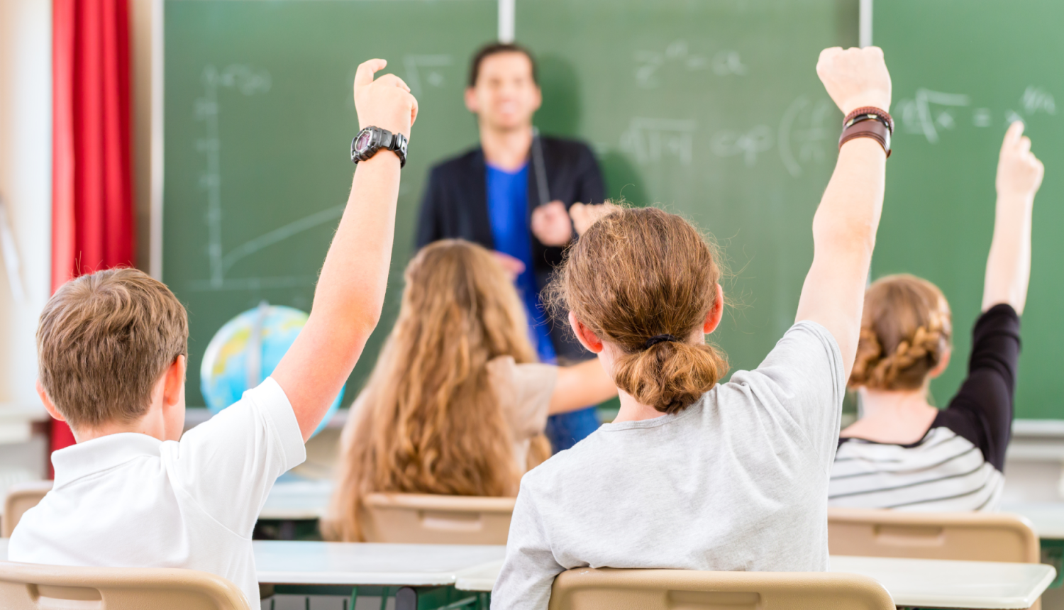 Skolēni pacēluši rokas, skolotājs stāv pie tāfeles