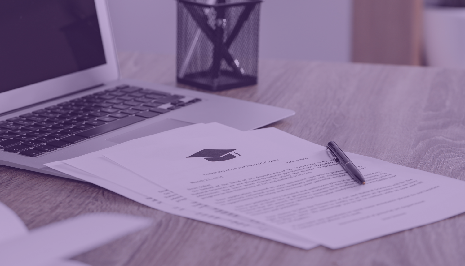 Uz galda stāv dators, dokumenti un pildspalva, uz dokumenta attēlota absolventa cepurītes ikona