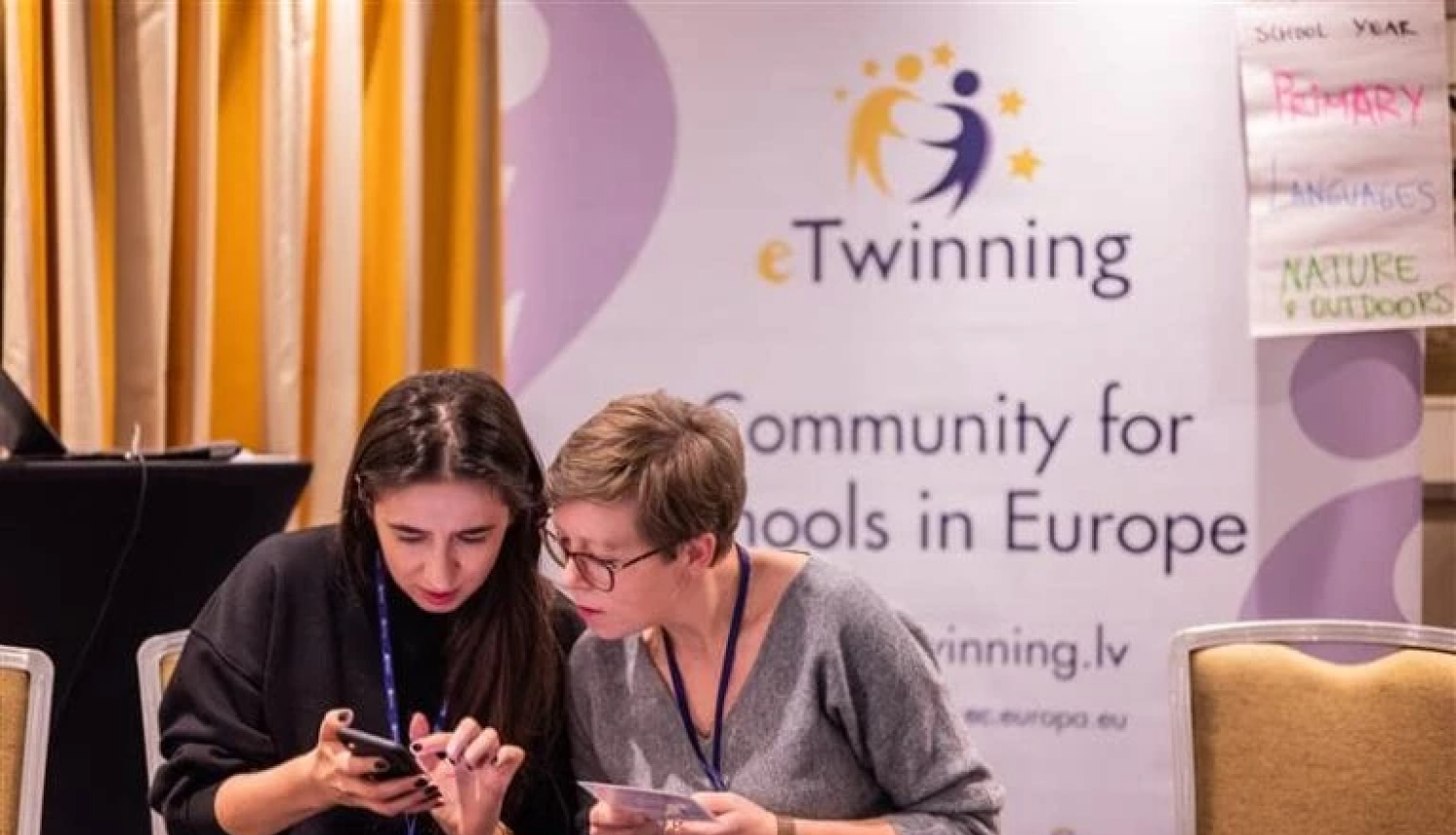 eTwinning baneris, priekšā tam sēž divas sievietes un pēta telefonu