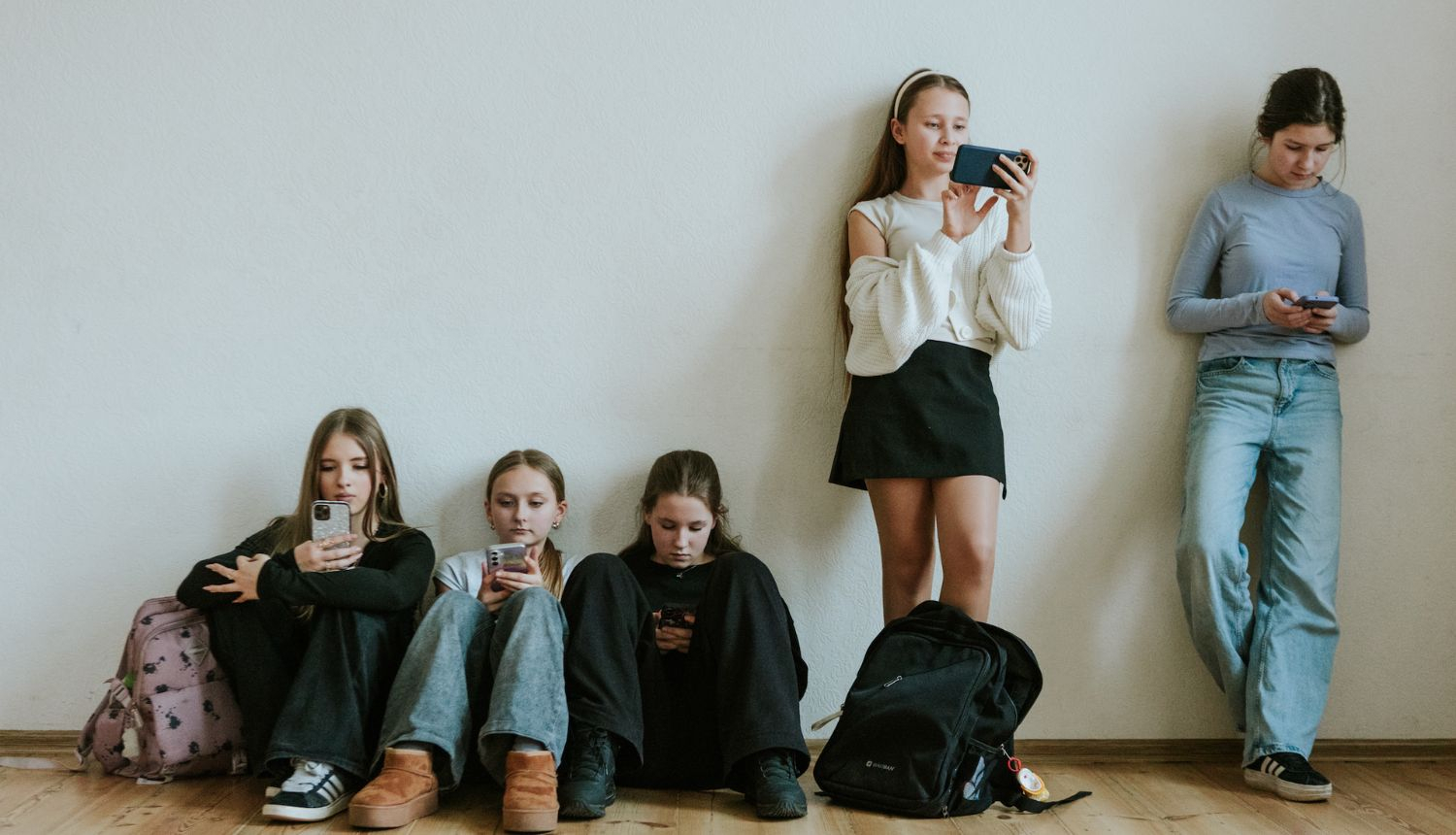 Skolēni atspiedušies pret sienu un apsēdušies uz zemes, pēta telefonus