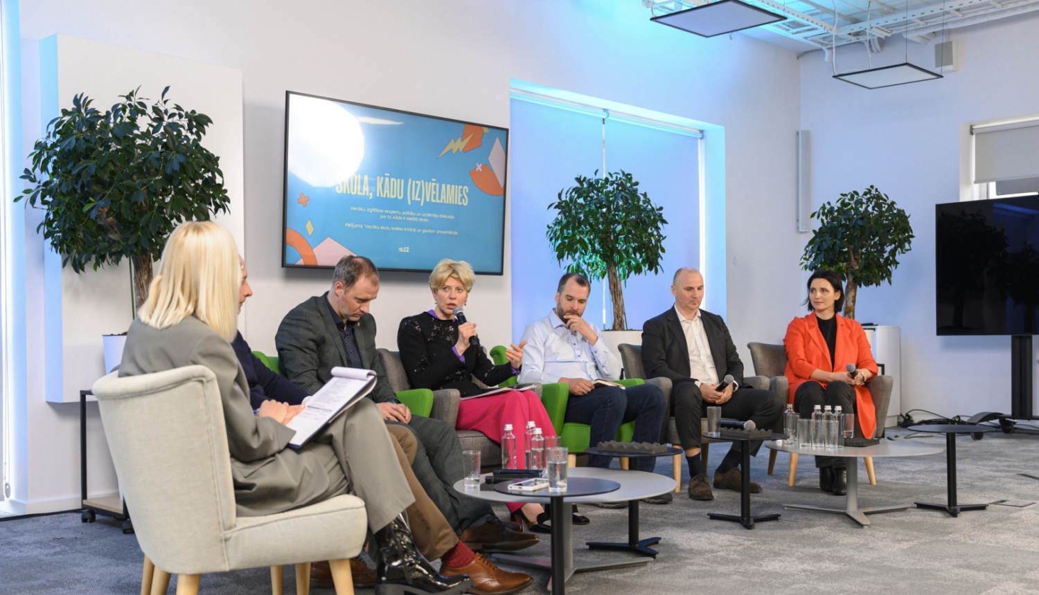 Izglītības un zinātnes ministre Anda Čakša piedalās diskusijā; septiņi cilvēki sēž pusaplī, ministre runā mikrofonā