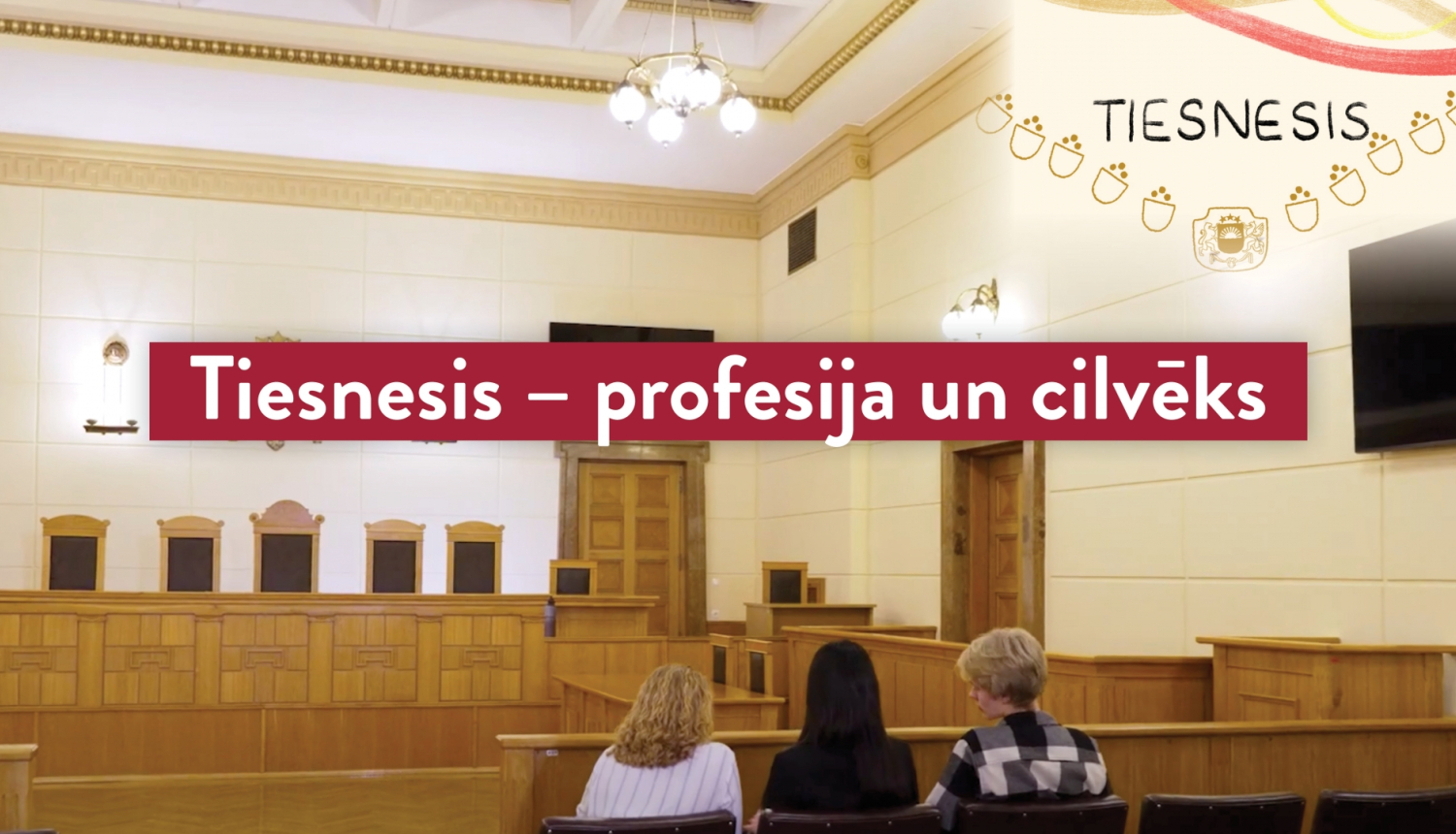 Attēlā redzama tiesas zāle un priekšplānā ir uzraksts baltiem burtiem uz sarkana fona "Tiesnesis - profesija un cilvēks"