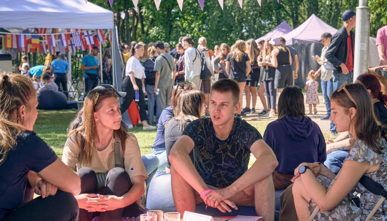 Jaunieši sēž un sarunājas, fonā daudz cilvēku un telts ar dažādu valstu karodziņiem