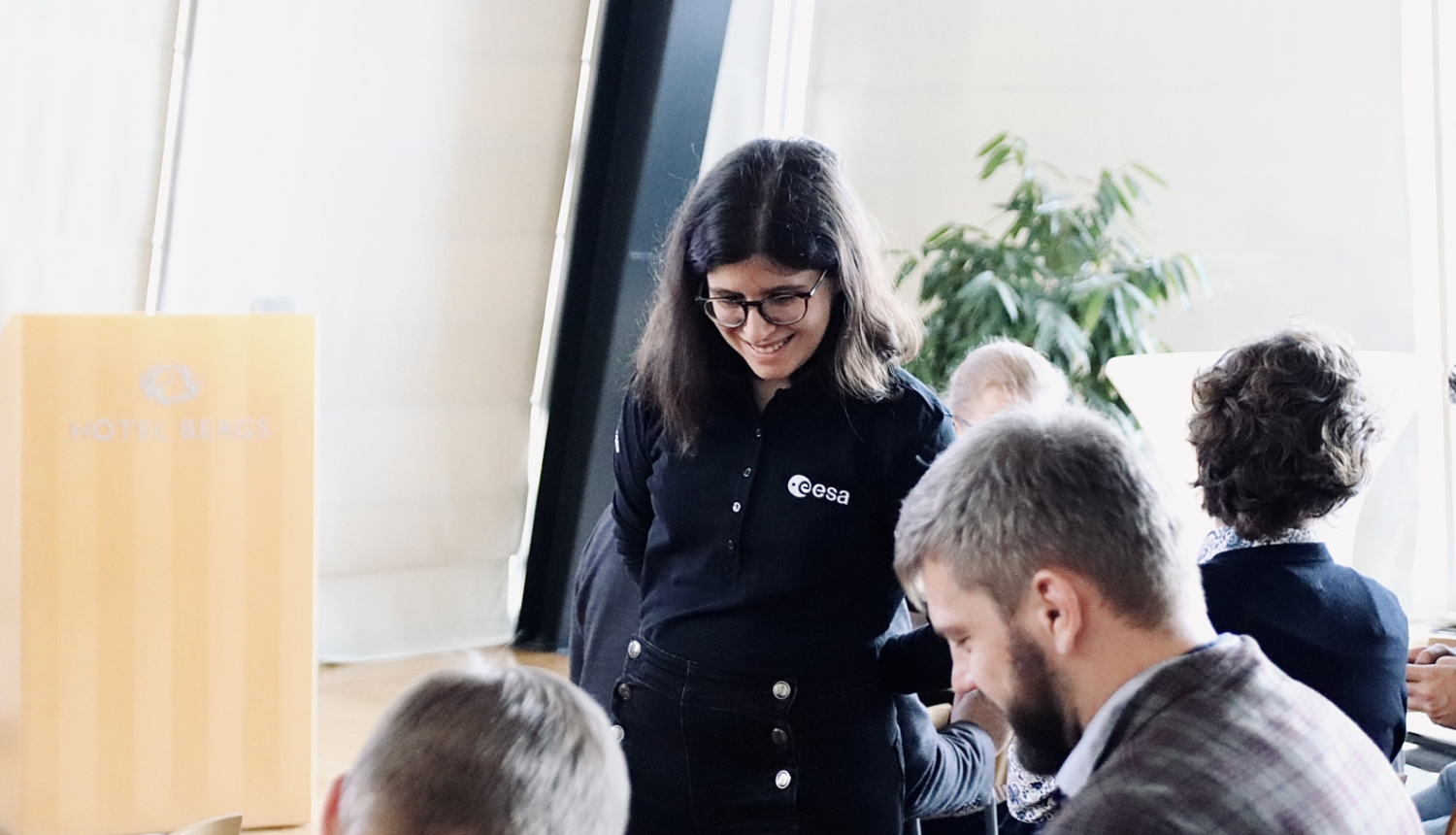 Smaidīga meitene stāv blakus galdam, kur sēž divi smaidīgi vīrieši. Notiek mācību process.