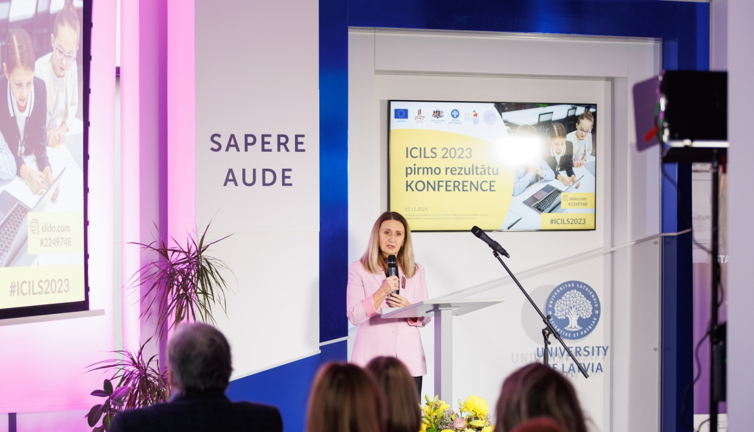 Inita Juhņēviča saka uzrunu konferencē, aizmugurē liels ekrāns ar uzrakstu "ICILS 2023 pirmo rezultātu prezentācija"
