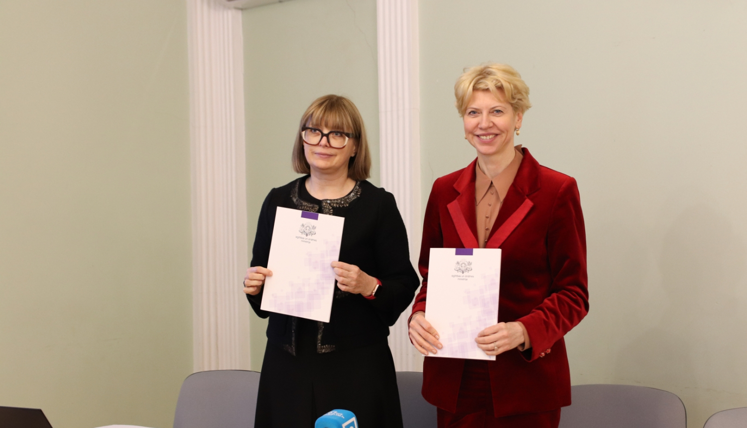 Attēlā redzama profesore Linda Daniela un izglītības un zinātnes ministre Anda Čakša, kas tur rokās mapes ar IZM logotipu