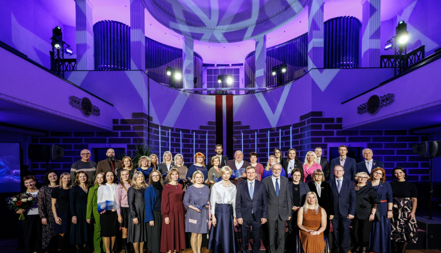 Violetās gaismās izgaismotā LU Lielajā aulā kopõgi stāv visi "Latvijas Gada skolotājs" balvas laureāti, izglītības un zinātnes ministre un LU rektors