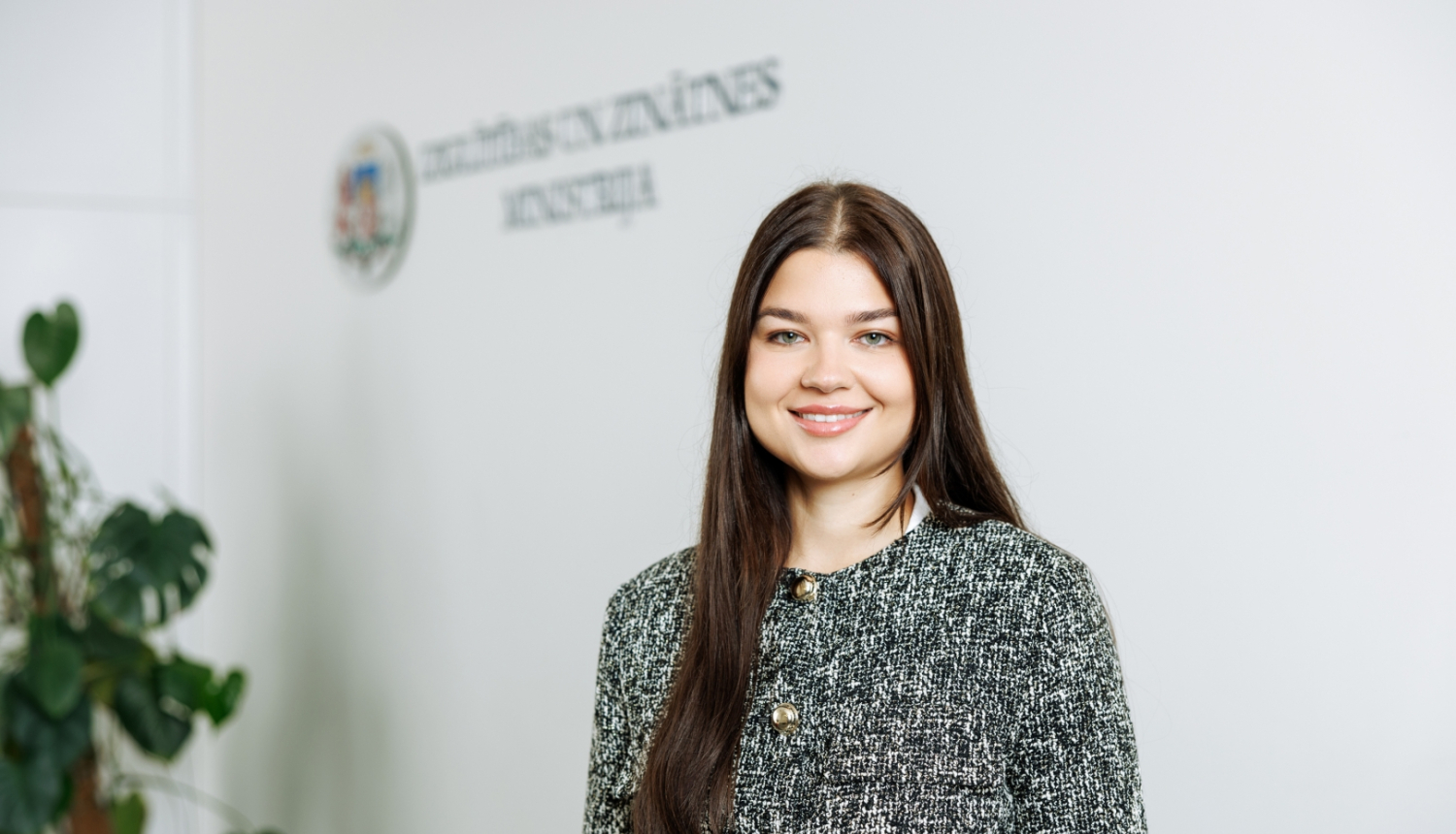 Silvija Amatniece stāv uz gaišas sienas fona. Uz sienas ir uzraksts "Izglītības un zinātnes ministrija"