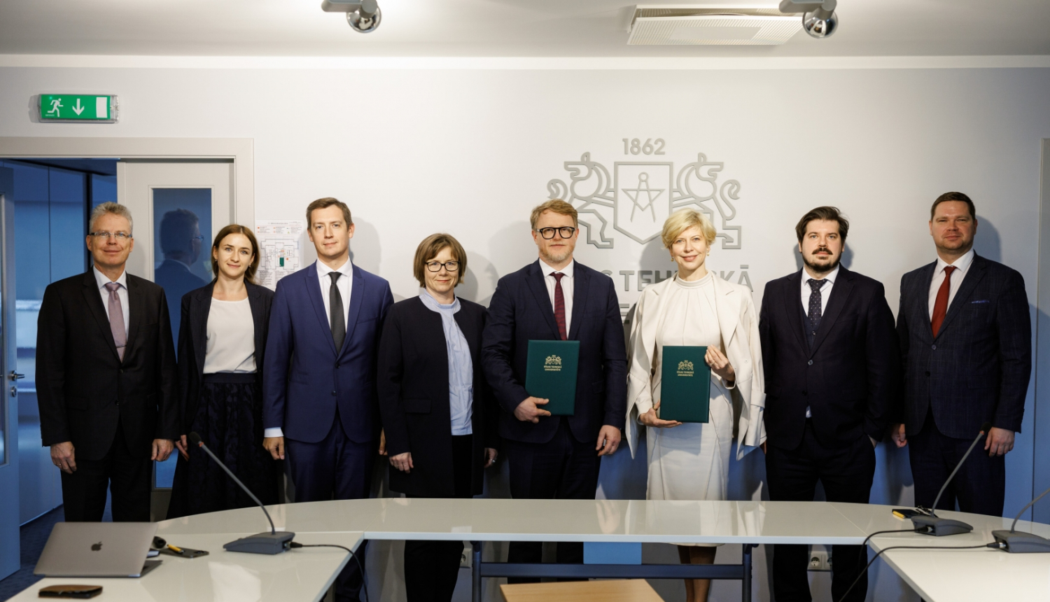 RTU pārstāvju un ministrijas pārstāvju kopbilde pie sienas, uz kuras redzams liels RTU logo