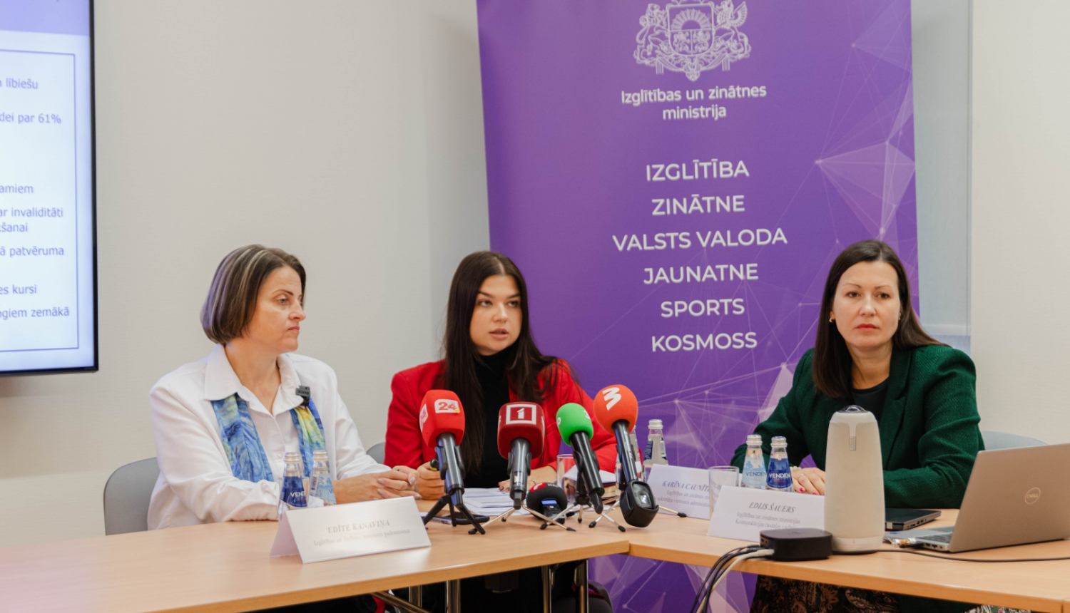 Pie galda sēž Edīte Kanaviņa, Silvija Amatniece un Karīna Caunīte-Orupe. Uz galda mediju mikrofoni, fonā uz violeta fona IZM logo un uzskaitītas zinātņu nozares: izglītība, zinātne, valsts valoda, jaunatne, sports, kosmoss