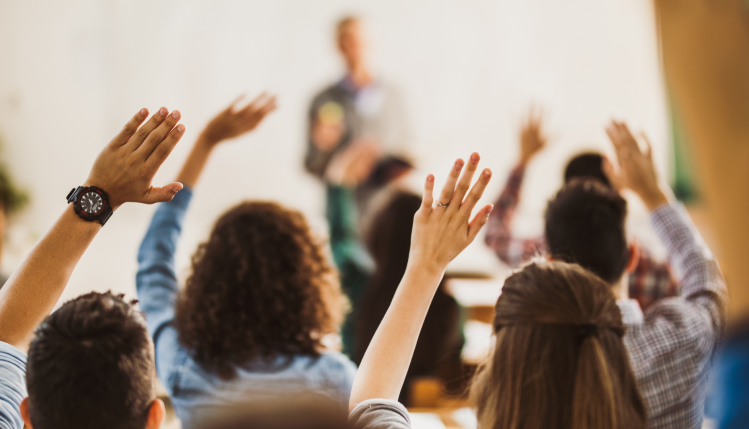 Bērni skolā ceļ roku, fonā stāv skolotājs