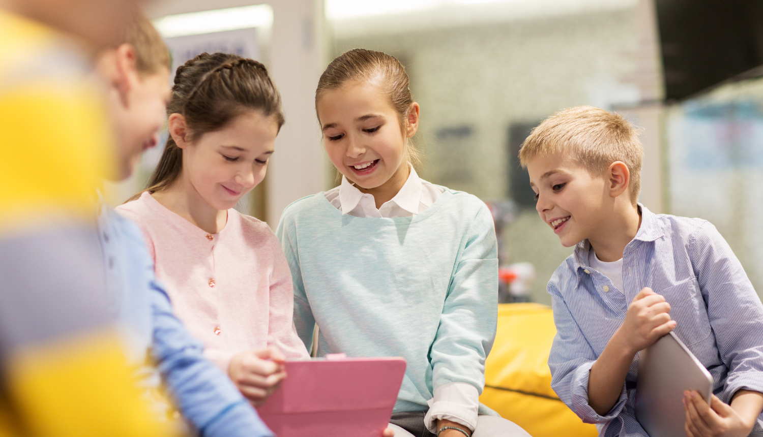 Pieci priecīgi bērni sēž kopā, skatās planšetē, fokusā divas meitenes un puika