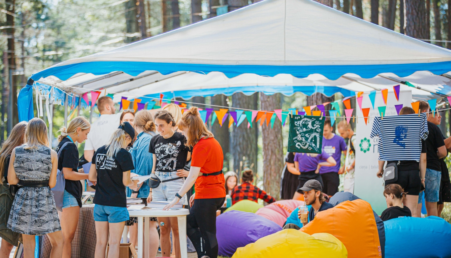 Jaunieši pulcējas pie telts. Ap telti sakārtas krāsainu karodziņu virtenes, blakus novietoti krāsaini sēžammaisi. 