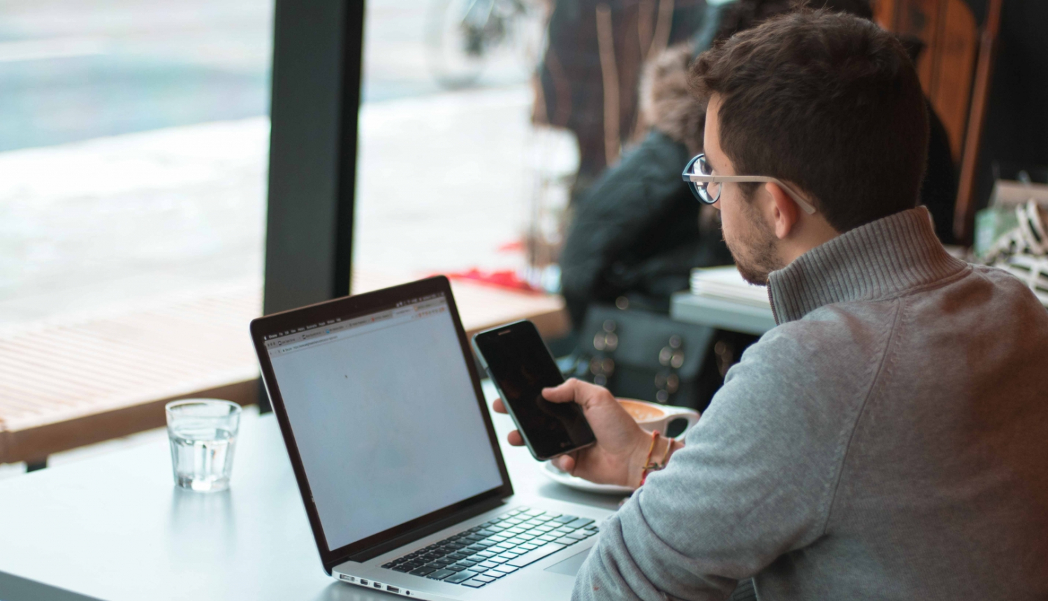 vīrietis ar telefonu rokās sēž pie galda, uz kura ir dators